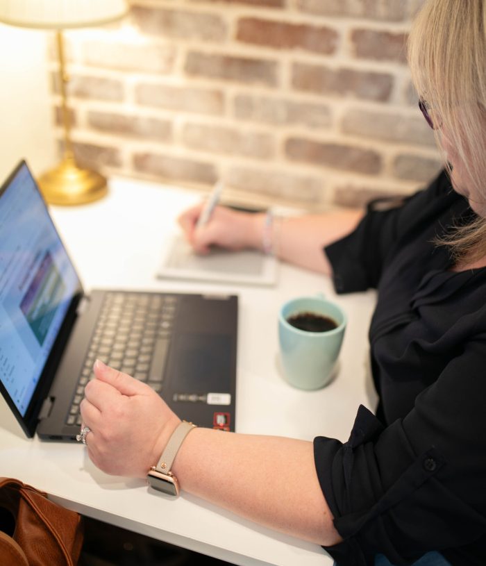 A blonde-haired woman works at a laptop, a blue cup of coffee next to her, with a light in the corner.