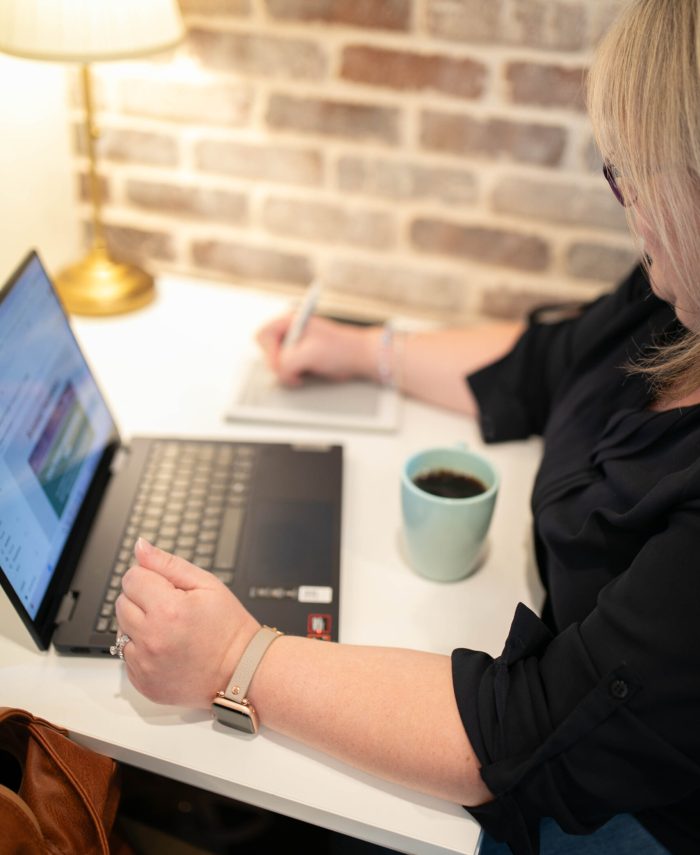 A blonde-haired woman works at a laptop, a blue cup of coffee next to her, with a light in the corner.