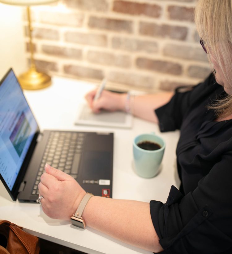 A blonde-haired woman works at a laptop, a blue cup of coffee next to her, with a light in the corner.