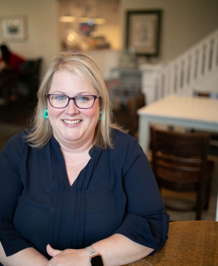 A blonde woman wearing a blue shirt, seafoam green earrings, and purple glasses, sits in a coffee shop.