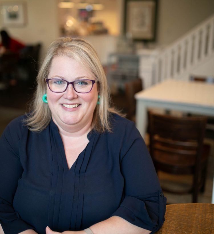 A blonde woman wearing a blue shirt, seafoam green earrings, and purple glasses, sits in a coffee shop.