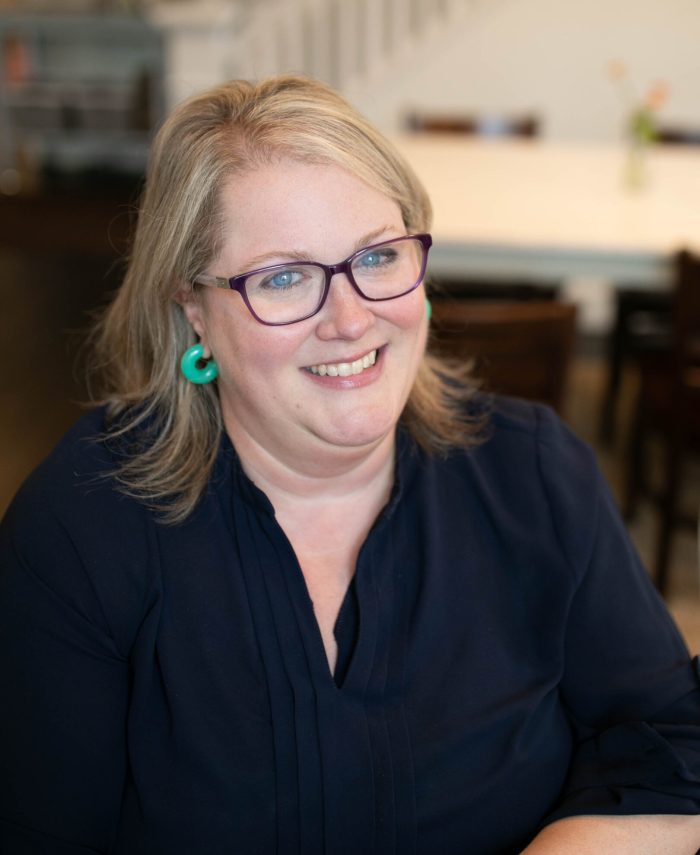 A blonde woman wearing a blue shirt, seafoam green earrings, and purple glasses sits in front of the camera.